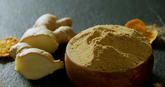 Close-up of fresh ginger root next to a wooden bowl filled with powdered ginger on dark surface. This image captures the natural texture and color of ginger, making it perfect for use in food blogs, cooking websites, health articles, and spice-related content.