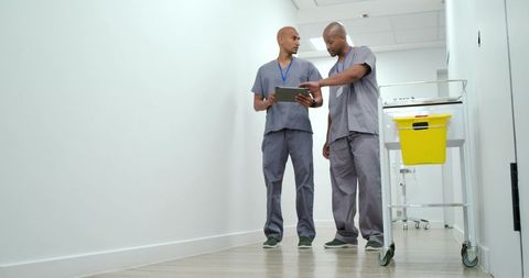 African american male clinicians reviewing tablet in hospital corridor with medical cart
