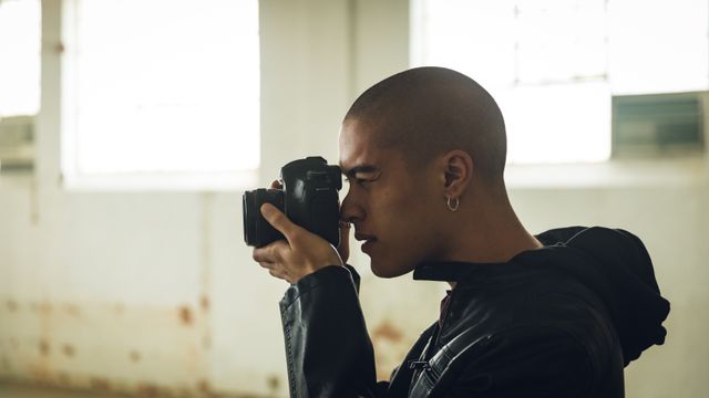 Side view of a hip young biracial man in an empty warehouse, taking photos with SLR camera, wearing black hoodie.