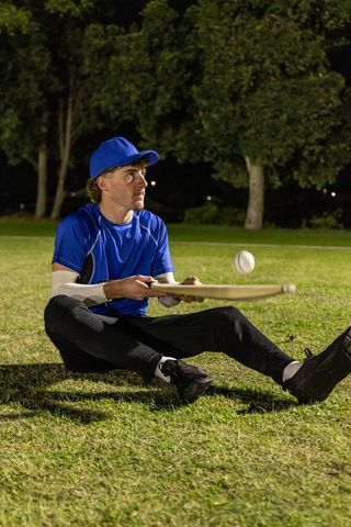Night practice baseball player sitting on grass catching ball with wooden bat under lights