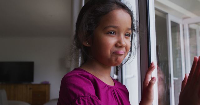 Young girl standing indoors, looking through glass door with a thoughtful expression, wearing purple shirt, natural light highlighting her face. Suitable for use in themes of childhood, contemplation, home, emotions, indoor activities.