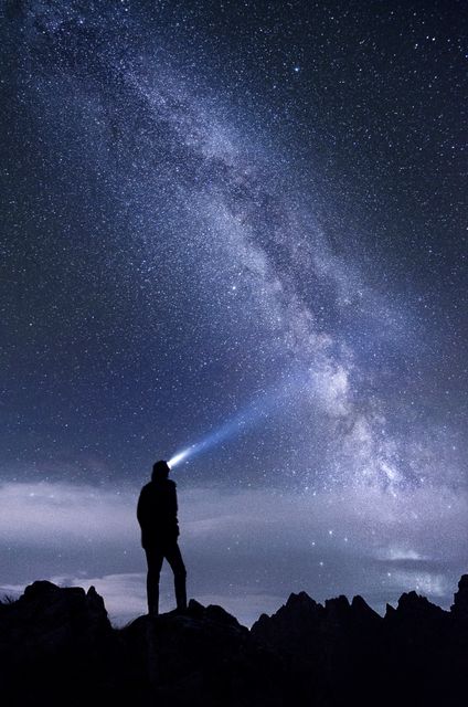 Silhouette of a person standing on rocky terrain at night, wearing a headlamp that illuminates the star-filled sky. The Milky Way galaxy is vividly visible, creating a scenic and awe-inspiring backdrop. This could be used in advertisements for outdoor gear, travel promotions, inspirational posters, space-themed content, or educational materials about astronomy.
