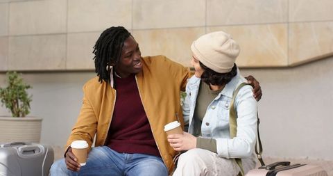 Diverse couple sharing coffee and smiles while waiting with suitcases at urban transit