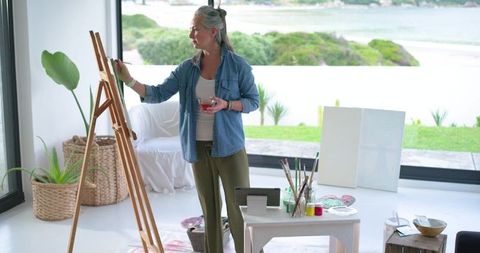 Mature woman painting in light-filled coastal studio holding brush and glass with easel