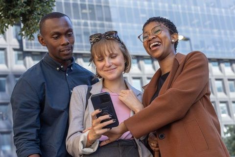Multiracial colleagues laughing, sharing smartphone, collaborating outside modern office