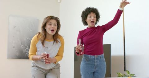 Diverse female friends reacting and cheering while playing living room game holding drinks