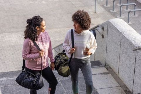 Two women walking up urban stairs with gym duffel bags and reusable water bottle, fitness friends
