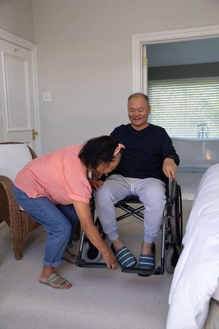Asian caregiver assisting senior man in wheelchair adjusting slippers at bedside homecare