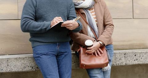 Mature couple leaning on city wall writing notebook holding coffee and leather purse