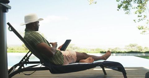 Middle-aged man relaxing on lounge chair by infinity pool using tablet, summer retreat