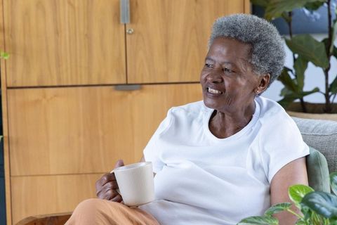 Senior african woman relaxing on sofa, holding coffee mug, smiling among indoor plants
