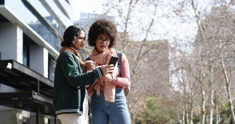 Friends sharing smartphone and coffee on urban sidewalk during autumn stroll