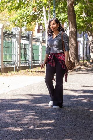 Relaxed urban woman walking along tree-lined path with plaid shirt tied and crossbody bag
