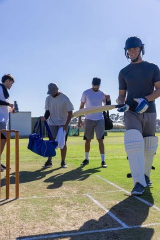 Cricket teammates walking onto pitch batter wearing helmet and pads under bright blue sky