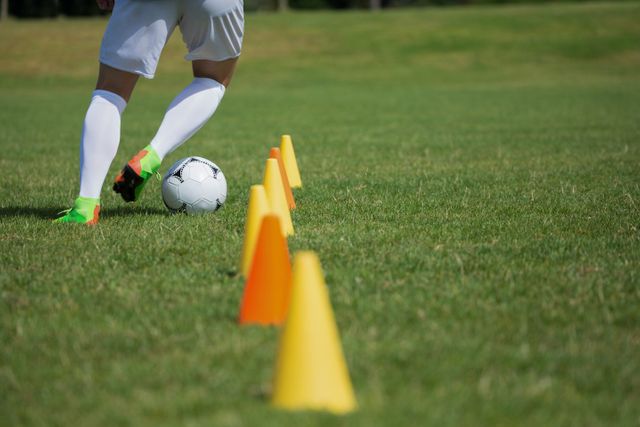 Image depicts a soccer player practicing dribbling through cones on a sunny day. Ideal for use in sports-related content, training manuals, coaching guides, and advertisements promoting soccer gear and training equipment.