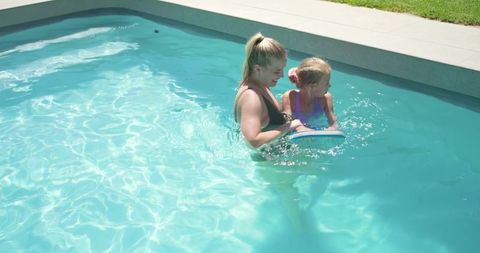 Caregiver teaching child swimming with kickboard in backyard pool, sunlit turquoise water