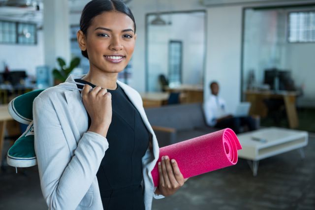 Businesswoman holding exercise mat and shoes, smiling in modern office. Ideal for promoting corporate wellness programs, work-life balance, and healthy lifestyle initiatives in the workplace. Suitable for use in articles, blogs, and advertisements related to employee wellness and fitness.