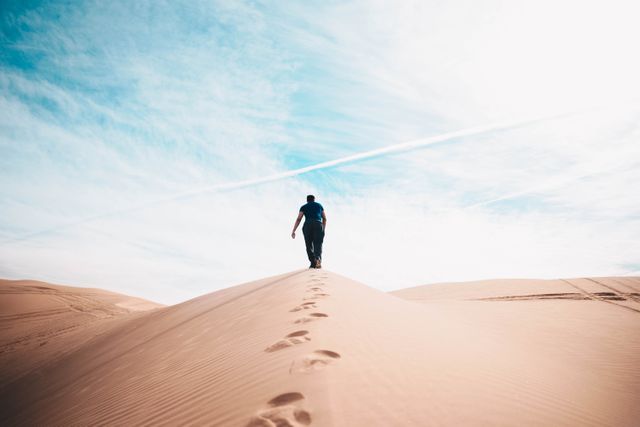 Depicts an individual walking on a sand dune in a vast desert under a bright sky with footprints trailing behind them. Perfect for illustrating themes of adventure, exploring, travel, solitude, or nature's vastness. Ideal for use in travel blogs, motivational materials, adventure magazines, or advertisements promoting outdoor activities.