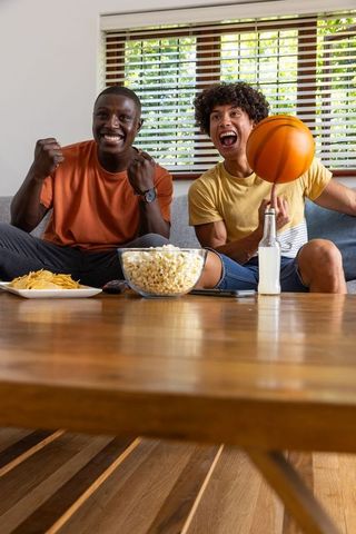 Diverse male friends cheering on sofa while spinning basketball during game night