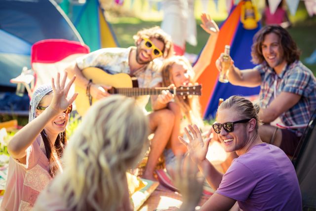 Group of friends enjoying a sunny day at a campsite, playing guitar, drinking, and waving. Ideal for use in advertisements, travel blogs, social media posts, and lifestyle articles promoting outdoor activities, friendship, and leisure.