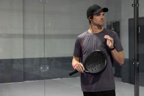 Padel player wearing cap holding black perforated paddle on indoor glass-walled court