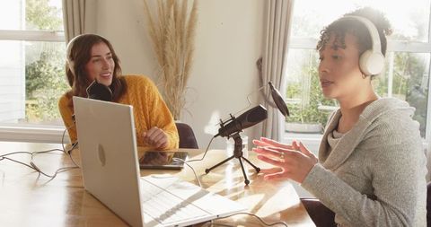 Two women co-hosting podcast in bright home studio with laptop, microphone and headphones