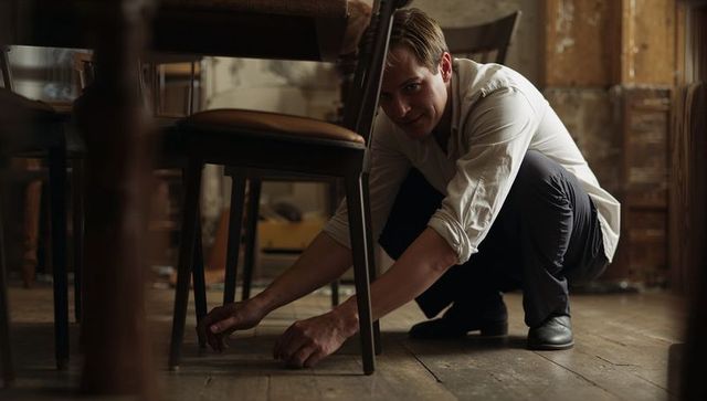 Crouching man reaching under chair in rustic dining room searching for small object