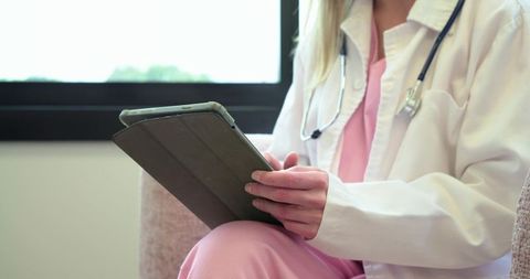 Adult female clinician using tablet for telemedicine consultation wearing white coat and stethoscope