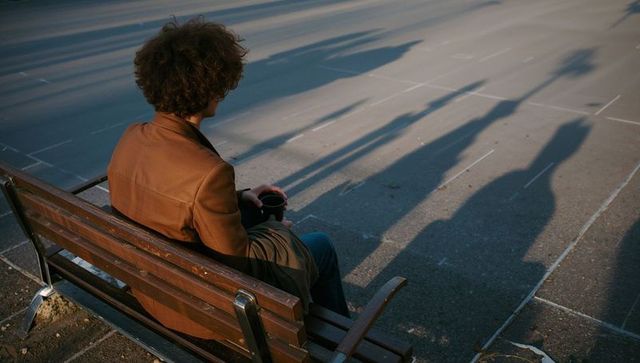 Solitary man sitting on bench under long shadows holding coffee cup in urban plaza