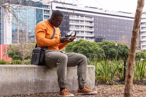 African american man adjusting headphones on bench in urban park with city skyline