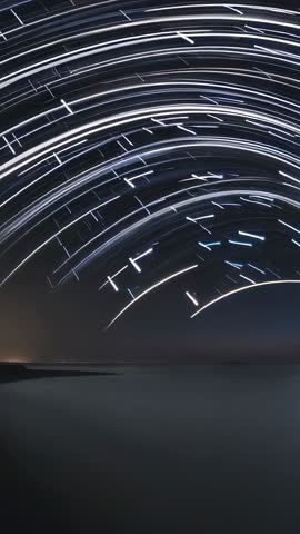 Vertical time-lapse star trails arcing over calm coastal water and shoreline at night