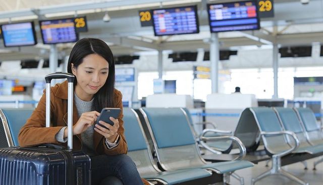 Solo traveler waiting at airport gate using smartphone with carry-on luggage