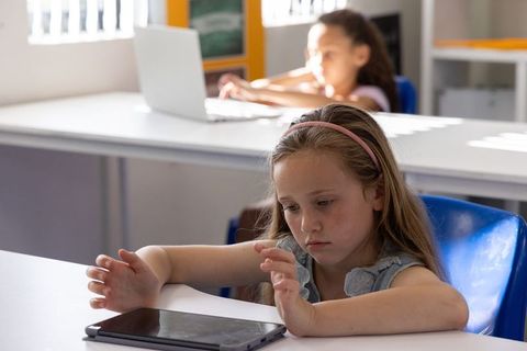 Elementary student using tablet in sunlit classroom while classmate works on laptop