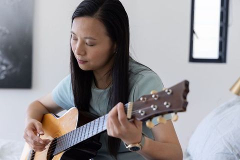 Mature asian woman sitting on bed playing acoustic guitar wearing light green top