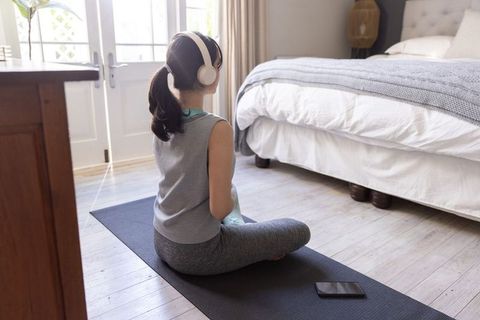 Woman practicing mindful meditation on yoga mat in bright bedroom wearing headphones