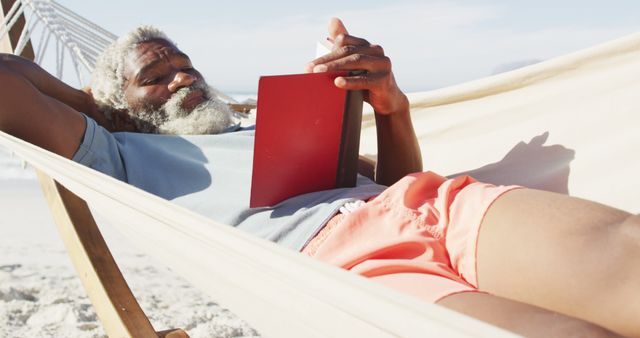 An elderly man is relaxing on a hammock by the beach, enjoying reading a book. The weather is clear and sunny, enhancing the serene atmosphere. This can be used for travel brochures, retirement planning materials, and advertisements focused on leisure and relaxation.
