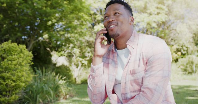 Man talking on phone while smiling, sitting in park with greenery. Great for themes of technology, communication, lifestyle, outdoor activities, relaxation, and happiness.