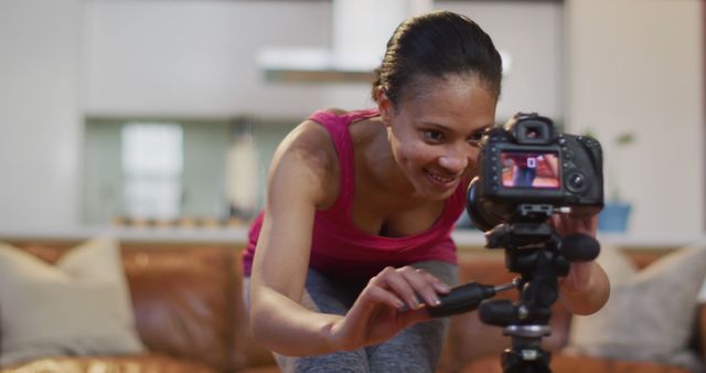 Young woman in casual clothing adjusting a digital camera mounted on a tripod. Ideal for concepts related to home video recording, vlogging, photography, creative professions, modern technology, and lifestyle. It can be used as a visual representation in articles, blogs, or advertisements promoting DIY video production, tutorial recording, and casual photography setups.