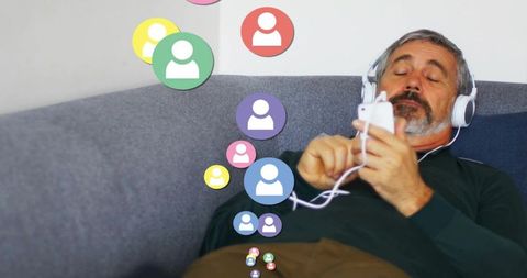 Mature man relaxing on sofa with wired headphones and smartphone browsing social avatars