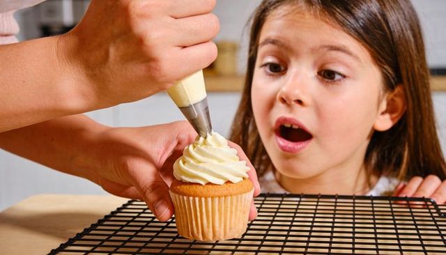 Adult Piping Vanilla Frosting on Cupcake While Girl Watches with Surprise in Home Kitchen