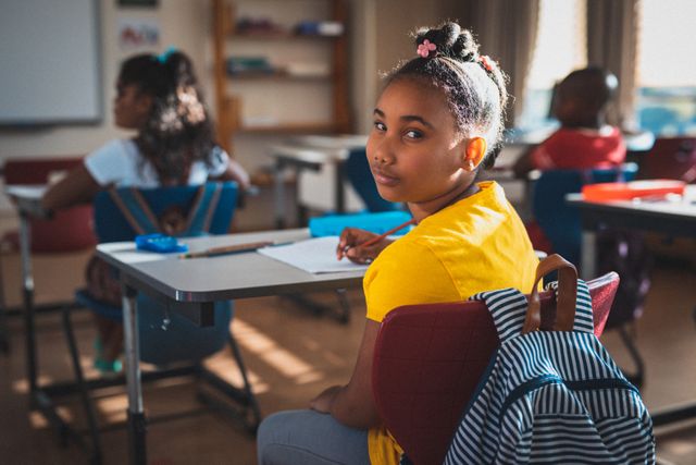 Young biracial schoolgirl sitting at desk in classroom, turning around to look at camera. Ideal for educational content, diversity in education, school advertisements, and childhood learning themes.