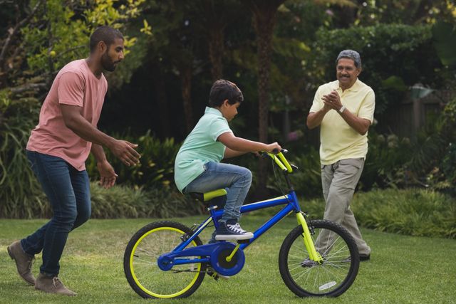 Father and grandfather encouraging young boy as he learns to ride a bicycle in a park. Ideal for use in family-oriented advertisements, parenting blogs, outdoor activity promotions, and educational materials about learning new skills and family support.