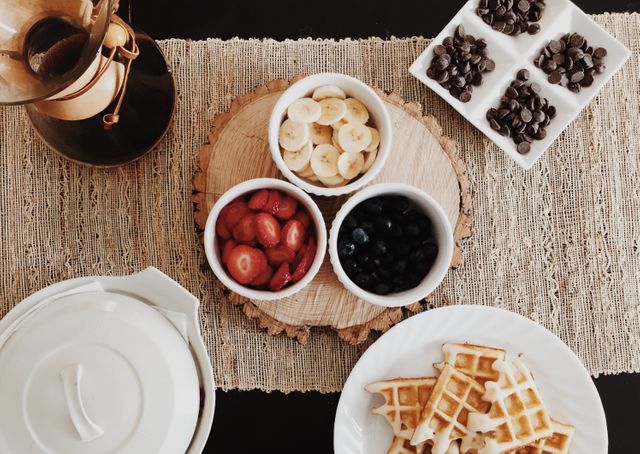 A beautifully presented breakfast spread featuring waffles, sliced strawberries, blueberries, banana slices, chocolate chips, and a pot of coffee. Perfect for use in food blogs, breakfast menu designs, healthy eating promotions, or lifestyle magazines focusing on morning routines or recipes.