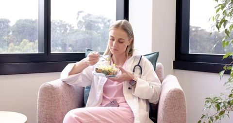 Nurse eating healthy salad during break in sunlit staff lounge armchair with stethoscope