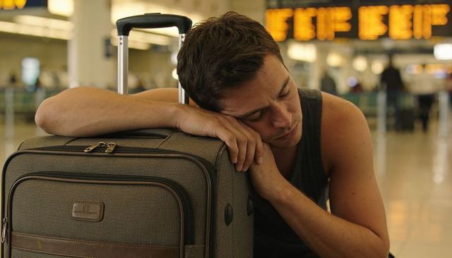 Resting male traveler leaning on wheeled suitcase at busy airport terminal, waiting, looking tired