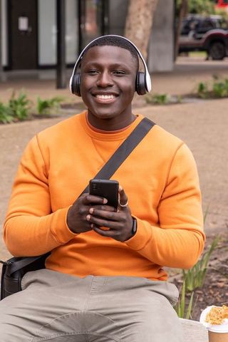 Smiling student wearing orange sweatshirt and headphones using smartphone on campus bench