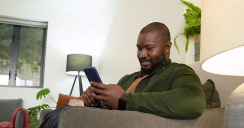 African American man relaxing on couch holding smartphone smiling in modern living room
