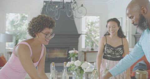 Friends arranging bottles and floral centerpiece on bright dining table for casual hosting