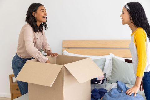 Mother and teenage daughter unpacking clothes from cardboard box on bed smiling, bonding