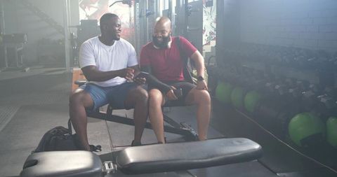 Two african american men relaxing after workout checking smartphone on gym bench